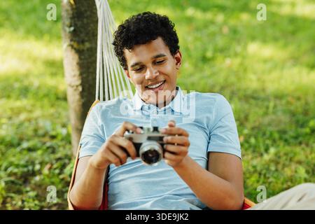 Giovane uomo sorridente che tiene in mano una macchina fotografica mentre sdraiato su un'amaca nel parco || modello rilasciato Foto Stock