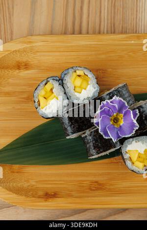 Panini di sushi pieni di pezzi di mango si trovano su una tavola di bambù. Un fiore viola aggiunge un tocco decorativo. Questo piatto presenta ingredienti freschi e skillfu Foto Stock