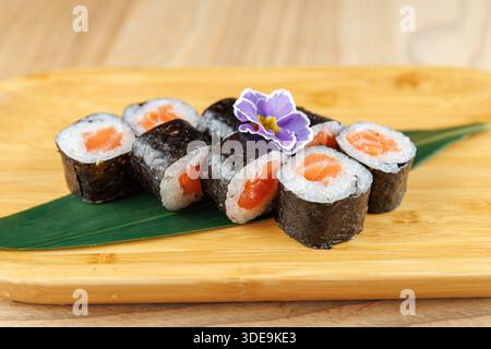 Gli involtini di sushi ripieni di salmone e riso sono ordinatamente disposti su una tavola di legno. Un fiore commestibile aggiunge un tocco di colore alla presentazione, creando un Foto Stock