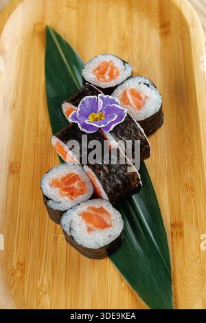 Una varietà di sushi ripieni di salmone fresco sono disposti su un piatto di bambù. Un fiore viola aggiunge un tocco colorato alla presentazione. Questo disp Foto Stock