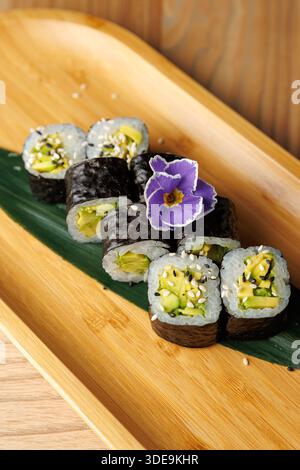Gli involtini di sushi ripieni di cetriolo e avocado sono disposti ordinatamente su una foglia verde. Un fiore aggiunge un tocco di colore e freschezza alla presentazione Foto Stock