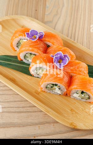 Gli involtini di sushi ripieni di cetriolo e formaggio spalmabile sono disposti ordinatamente su un piatto di legno. Le fette di salmone arancione brillante coprono gli involtini, con fiori per Foto Stock