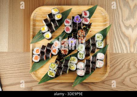 Vari sushi roll sono disposti ordinatamente su un piatto di bambù. Il display include rotoli colorati con diversi ripieni, decorati con foglie verdi Foto Stock