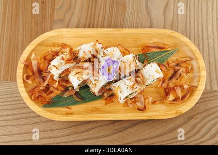 Un piatto di legno mostra sushi Rolls ricoperti di fiocchi di bonito e un piccolo fiore. L'ambiente è luminoso, mettendo in risalto gli ingredienti freschi e la cura Foto Stock