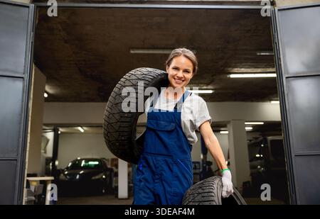 Felice giovane meccanico di auto che trasporta pneumatici in un'officina di riparazione auto Foto Stock
