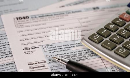 Una persona lavora su 1040 moduli fiscali utilizzando una calcolatrice e una penna in un ufficio domestico. Il documento descrive in dettaglio lo stato di archiviazione, i dipendenti e le risorse digitali. Foto Stock