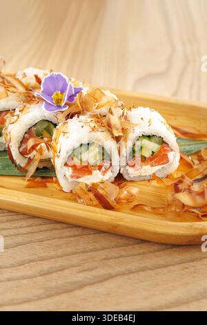 Gli involtini di sushi freschi sono disposti ordinatamente su un piatto di legno. I panini contengono cetriolo e salmone, ricoperti di fiocchi di bonito e guarniti con un fiore. Foto Stock