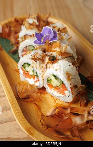 Panini di sushi con cetriolo, salmone e formaggio spalmabile serviti su un piatto di legno. Ricoperto di fiocchi di bonito e decorato con un fiore viola. Perfetto Foto Stock