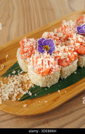 Un piatto di legno offre sushi Rolls ricoperti di gamberi e ingredienti grattugiati. Un fiore commestibile aggiunge colore al piatto. L'impostazione evidenzia Foto Stock
