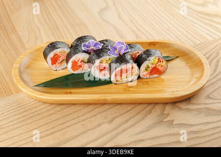 Panini di sushi ripieni di pesce fresco e verdure seduti su un piatto di bambù. Il piatto comprende fiori decorativi e una foglia verde. L'impostazione è semplice Foto Stock