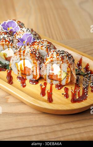 Una selezione di sushi roll è disposta su un piatto di legno. I panini sono pieni di ingredienti e ricoperti di salsa e piccoli fiori per la decorazione Foto Stock