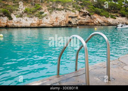 Scala metallica per piscine che conduce all'acqua di mare turchese nella baia di Cala Santanyi a Maiorca, Spagna. Primo piano di acque cristalline ondulate e scogliere sopra la S. Foto Stock