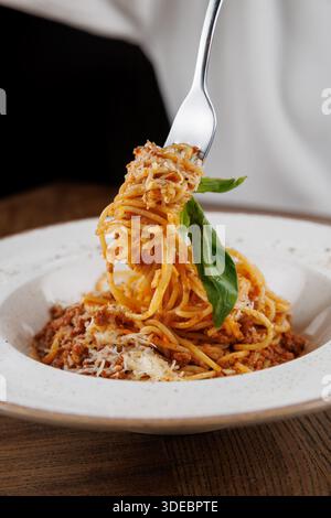 Una forchetta solleva gli spaghetti ricoperti di salsa di carne da una ciotola bianca. Il basilico verde si trova sopra e il formaggio grattugiato è cosparso sul piatto. Lo sfondo Foto Stock