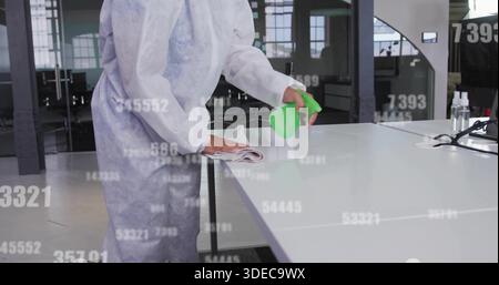 Donna di pulizia in tuta bianca che spruzza e pulisce la scrivania in ufficio, con flacone verde e igienizzatore Foto Stock