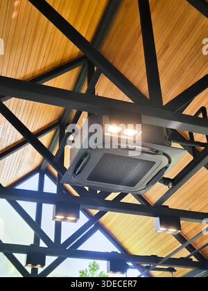 Condizionatore d'aria di tipo a cassetta montato su una struttura industriale a soffitto con travi a capriata in acciaio nero e tetto in legno che creano un'atmosfera moderna Foto Stock