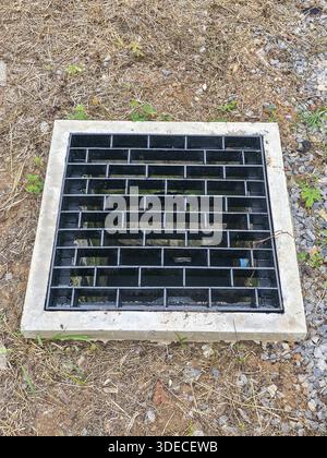 Coperchio quadrato della griglia di scarico in metallo nero con bordo in calcestruzzo su terreno sporco che mostra un design industriale robusto e un infrastrutturatore funzionale del sistema di drenaggio Foto Stock