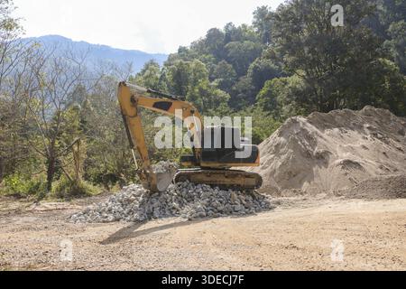 La macchina da scavo gialla lavora sodo con un potente movimento di scavo su un mucchio di rocce contro il bellissimo paesaggio verde della foresta montana durante un'intensa attività di scavo Foto Stock