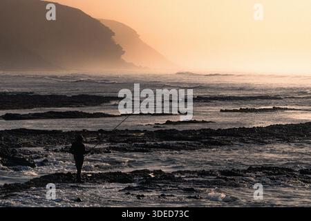 Silhouette di Fisherman sulla costa rocciosa all'alba con scogliere nebbiose Foto Stock