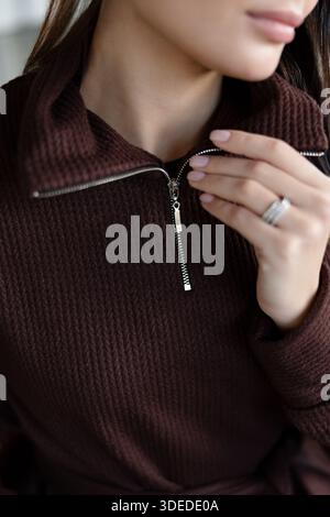 Foto dettagliata di zip metallica su camicia a manica lunga testurizzata marrone. Mano femminile con colletto a contatto con anello di un elegante indumento a coste. Concentrati sul tessuto Foto Stock