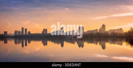 Tramonto sull'argine di Obolon a Kiev con moderno skyline residenziale che si riflette nelle calme acque del fiume, nelle spettacolari nuvole e nella luce calda Foto Stock