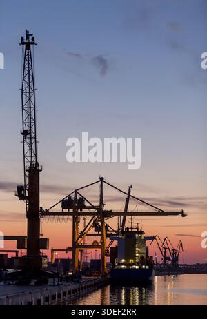 Sagoma di una nave portacontainer ormeggiata accanto alle gru portacontainer nel porto al tramonto, Klaipeda, Lituania Foto Stock