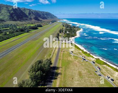 Vista aerea del vivace campo d'aviazione verde che contrasta con l'oceano turchese che incontra la spiaggia sabbiosa, con le montagne di Waianae sullo sfondo, Foto Stock