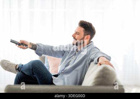 Uomo rilassato che passa da un canale all'altro con telecomando mentre guardi la tv a casa, rilassati sul divano, vista laterale Foto Stock