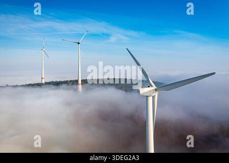 Vista aerea delle turbine eoliche che attraversano un mare di nebbia, le loro forme bianche si scontrano con il cielo blu profondo e la foresta verde scuro sottostante, Groß-um Foto Stock