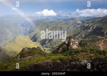 Vista ad alto angolo di un rifugio vicino a Pico Ruivo sull'isola di Madeira, in Portogallo, con un debole arcobaleno che si innalza sulla profonda valle verde Foto Stock