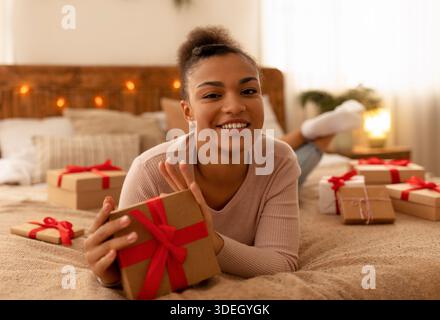 Felice giovane afroamericana signora sdraiata a letto e tenendo in mano la scatola regalo di Natale, sorridendo alla macchina fotografica Foto Stock