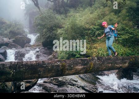 Giovane donna con zaino e pali da trekking che attraversa il ponte di legno vicino alla potente cascata del fiume di montagna durante il trekking nel Parco Nazionale di Makalu Barun Foto Stock