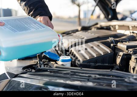 Uomo che versa liquido lavavetri blu dal contenitore in plastica nel serbatoio sotto il cofano della vettura aperto nelle giornate di sole. Concetto di manutenzione del veicolo, s Foto Stock