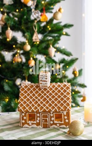 Casa di pan di zenzero con tetto a reticolo e finestre di caramello luminose di fronte all'albero di Natale decorato con ornamenti dorati e luci fiabesche. Concetto o Foto Stock