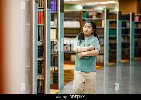 Studentessa asiatica che si muove attraverso gli scaffali della biblioteca con il libro in braccio. L'aspetto evidenzia l'indipendenza l'attenzione la curiosità l'istruzione la crescita la scuola positiva Foto Stock