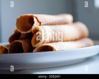 Coni di wafer arrotolati impilati su una piastra di ceramica bianca, natura morta di alimenti ravvicinata con profondità di campo ridotta. Foto Stock