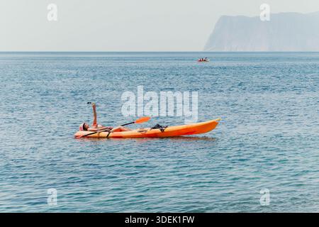 Kayak sulla costa del mare persona che si rilassa in un kayak arancione brillante su un mare calmo con una scogliera lontana sullo sfondo. Foto Stock