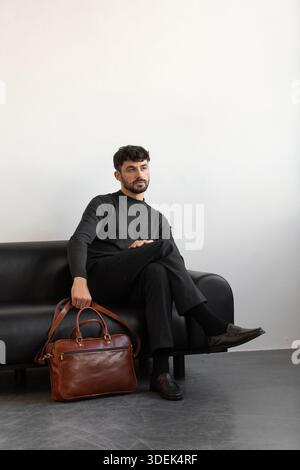 Un modello professionale maschile in maglieria a carbone e pantaloni sartoriali si trova su un divano in pelle nera con una valigetta in pelle marrone premium Foto Stock