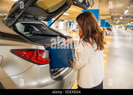 Donna che carica borse della spesa nel bagagliaio dell'auto nel parcheggio pubblico dopo gli acquisti. Routine di shopping quotidiana, trasporto personale e partenza Foto Stock