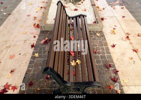 Foglie autunnali sparse intorno a una panchina di legno su una passerella acciottolata ad Abano Terme, Italia Foto Stock
