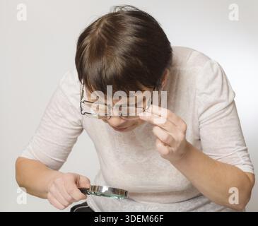 Donna anziana guarda qualcosa attraverso due bicchieri e una lente di ingrandimento Foto Stock