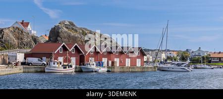 Un pittoresco porto svedese sull'isola di Tjorn, caratterizzato da boatouses in legno rosso, barche ormeggiate e uno sfondo roccioso. Acque blu calme, cielo limpido e un pe Foto Stock