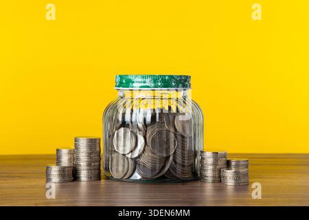 Monete che riempiono un vaso di vetro e impilate su un tavolo di legno su sfondo giallo Foto Stock