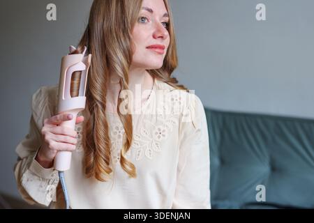 Donna che si prende cura di sé mentre arriccia i capelli a casa con una luce soffusa e un'atmosfera tranquilla Foto Stock