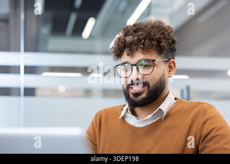 Giovane uomo barbuto con capelli ricci e occhiali che si concentra sul suo computer portatile mentre lavora in un ambiente d'ufficio contemporaneo, mostrando dedizione e concentrazione Foto Stock