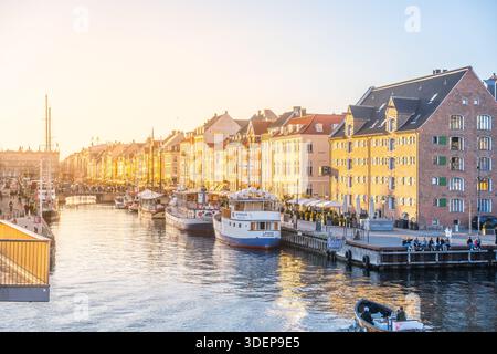 Nyhavn mostra colori vivaci mentre il sole tramonta sull'iconico porto. Le barche fiancheggiano l'acqua mentre le persone si godono il caldo bagliore della serata in Danimarca, un'incantevole posizione. Foto Stock