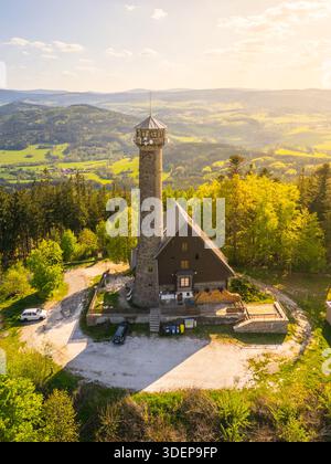 Il drone cattura una vista aerea della torre di osservazione di Svatobor circondata da lussureggiante vegetazione e colline ondulate in Cechia al tramonto, mostrando la bellezza del paesaggio e dell'architettura. Foto Stock