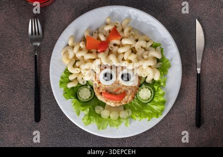 Cibo divertente per bambini - una ragazza sorridente di tacchino, cavatappi di pasta, olive, pepe, uovo e cetriolo su piatto bianco, vista dall'alto Foto Stock