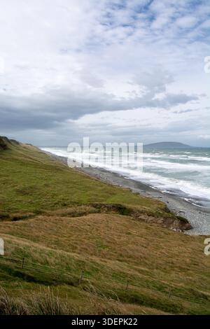 Un paesaggio costiero panoramico caratterizzato da colline verdi ondulate, una costa rocciosa e turbolente onde oceaniche sotto un cielo nuvoloso. Foto Stock