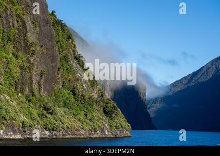 Un tranquillo paesaggio caratterizzato da ripide e lussureggianti scogliere verdi che si innalzano da un tranquillo corso d'acqua blu, con nebbia che avvolge dolcemente le cime delle montagne sotto un cle Foto Stock