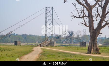 Un semplice ponte sospeso si estende su una pianura pianeggiante e erbosa punteggiata di alberi sparsi, creando un paesaggio sereno e aperto. Foto Stock
